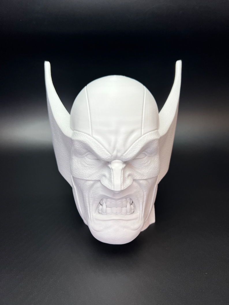 May include: A white, 3D-printed bust of a character's head, featuring a stern expression with furrowed brows and bared teeth. The head has pointed, wing-like structures on either side, and a mask covering the eyes.
