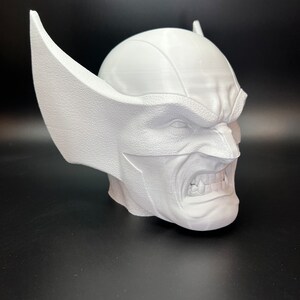 May include: A white 3D-printed bust of a character with a stern expression, featuring pointed ears and a textured surface. The bust is set against a dark background, showcasing the detailed design of the superhero's face.