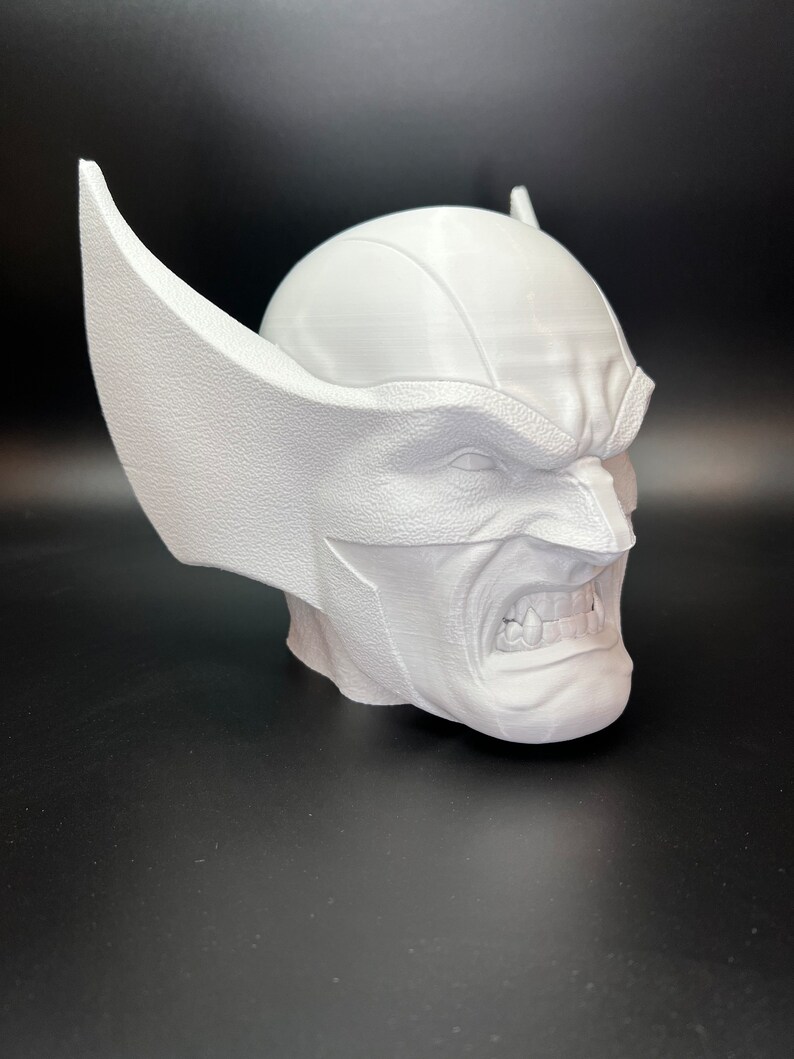 May include: A white 3D-printed bust of a character with a stern expression, featuring pointed ears and a textured surface. The bust is set against a dark background, showcasing the detailed design of the superhero's face.