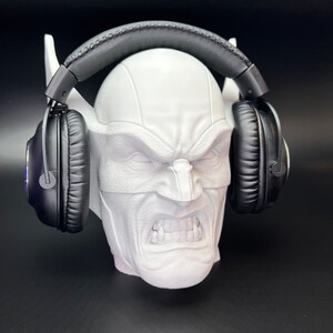 May include: A white, sculpted head with a stern expression, holding a pair of black headphones. The head has pointed ears and a textured surface. The headphones have black earcups and a padded headband.