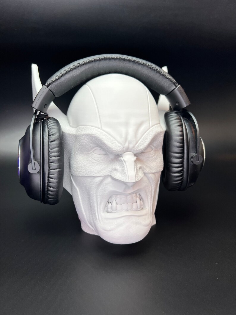 May include: A white, sculpted head with a stern expression, holding a pair of black headphones. The head has pointed ears and a textured surface. The headphones have black earcups and a padded headband.