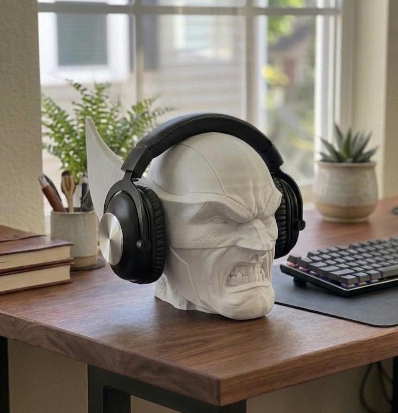 May include: A white, sculpted head with an angry expression, designed to hold headphones. The headphones are black with silver accents. The head is on a wooden desk, with a keyboard and plants in the background.