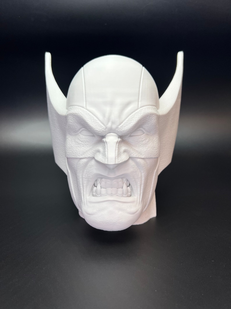 May include: A white, 3D-printed bust of a character's head, featuring a stern expression with furrowed brows and bared teeth. The head has pointed ear-like protrusions on each side.