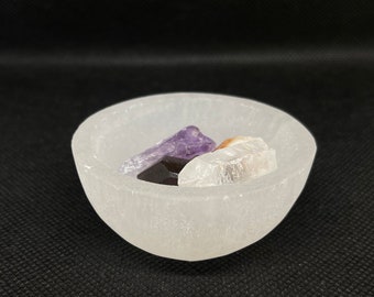 Könnte beinhalten: Eine weiße Selenit Schale mit einem violetten Amethyst, einem klaren Quarzkristall und einem schwarzen Obsidian Kristall.