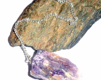Pode incluir: Um pêndulo de cristal de ametista bruto com uma corrente prateada. O cristal roxo é grosseiramente esculpido e de formato irregular. A corrente é presa ao topo do cristal e é colocada sobre uma pedra cinza.