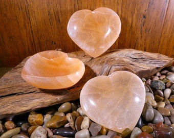 May include: Three heart-shaped bowls made of translucent orange alabaster. The bowls are arranged on a piece of driftwood with small stones surrounding them.