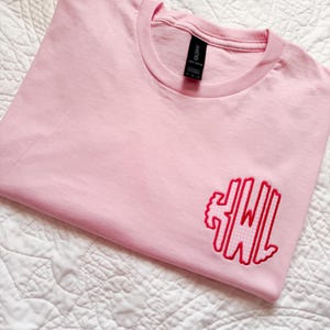May include: A light pink t-shirt with a crew neck and short sleeves. The shirt features a red embroidered monogram on the front. The shirt is folded neatly on a white quilted surface.