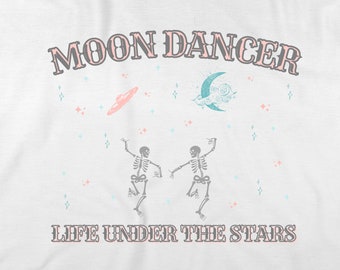 May include: White t-shirt with a pink and gray graphic design. The design features the text "Moon Dancer" and "Life Under The Stars" with two skeletons dancing under a crescent moon and stars.