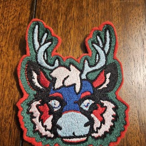 Könnte beinhalten: Gestickter Aufnäher mit einem stilisierten Hirschkopf-Design. Der Aufnäher hat einen roten Rand, einen grünen Hintergrund und ein Hirschgesicht mit blauen, roten, schwarzen und weißen Akzenten. Das Geweih ist hellblau.