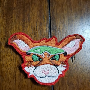 May include: Embroidered patch of a cartoon orange and white fox wearing a green visor. The fox has a pink nose and white whiskers.