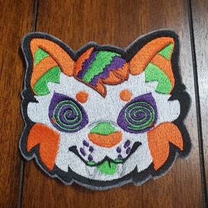 Może przedstawiać: Naszywka haftowana przedstawiająca białą kreskówkową postać zwierzęcia z pomarańczowymi, zielonymi i fioletowymi detalami. Zwierzę ma szeroki uśmiech z zielonym językiem i wirującymi zielonymi oczami.
