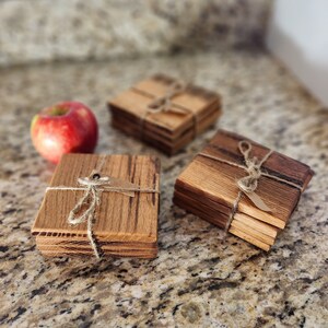 Puede incluir: Tres juegos de posavasos de madera, cada juego apilado y atado con cuerda. Los posavasos son de madera oscura con un patrón de veta natural. Los posavasos están sobre una encimera moteada.