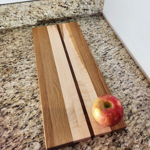 May include: A rectangular wooden cutting board with a light brown, dark brown, and white wood grain pattern. The board has a red apple on top of it.