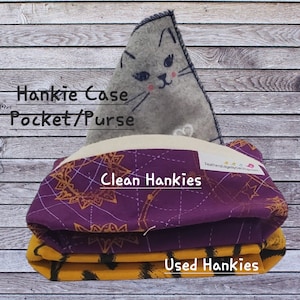 May include: A purple and gold fabric hankie case with a cat face on the top. The case has two compartments, one for clean hankies and one for used hankies. The text "Hankie Case Pocket/Purse" is written on the front of the case.