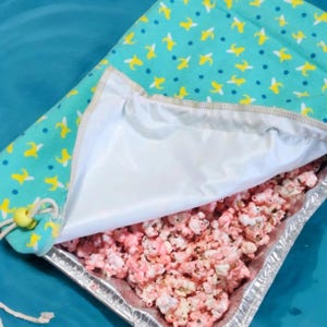 May include: A white fabric bag with a blue and yellow banana print pattern is partially open, revealing a silver pan filled with pink popcorn.