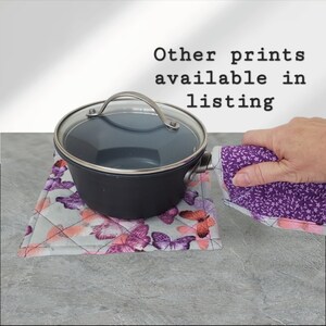 May include: A purple and gray quilted pot holder with a butterfly print. A black pot with a glass lid sits on top of the pot holder. A hand is holding a purple and white floral print pot holder.