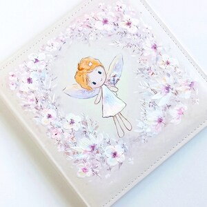 May include: A white leather journal with a hand-painted design of a fairy with blonde hair and a white dress surrounded by a wreath of white flowers.