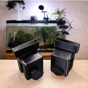 May include: Two black, 3D-printed aquarium tank clips. Each clip has a rectangular top piece, a square body, and a hexagonal screw. The clips are designed to hold aquarium equipment in place.