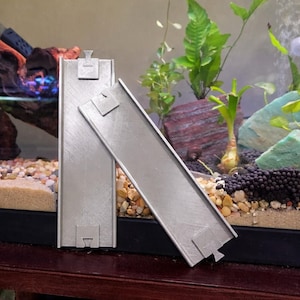 May include: Two silver-colored, rectangular aquarium accessories with a textured surface. Each has a small, square tab at the top and a notched design at the bottom. The items are positioned in front of an aquarium with plants and rocks.