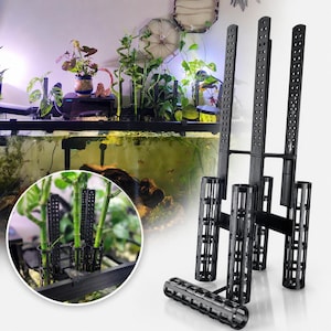May include: A black plastic aquarium plant support system featuring three vertical perforated stakes connected to cylindrical mesh baskets. The structure is designed to hold aquatic plant stems upright within a tank, with a circular inset showing the product in use with green plant stalks.