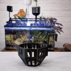 May include: A black, geometric aquarium plant holder with a rectangular hook. The holder has a series of vertical slots and is designed to hang inside an aquarium. The background features an aquarium with plants and a light.