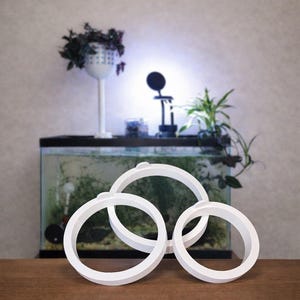 May include: Three white, circular, decorative objects with a raised, rounded design. The objects are arranged on a wooden surface in front of an aquarium and plants. The background includes a white plant stand and a small lamp.