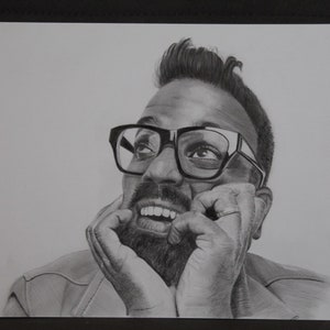 Peut inclure: Dessin au crayon d'un homme barbu portant des lunettes et regardant vers le haut avec un sourire. Le dessin est en noir et blanc, avec des ombres et des reflets détaillés. Les mains de l'homme reposent sur ses joues.