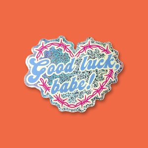 May include: A holographic sticker in the shape of a heart with pink barbed wire around the edge. The sticker says "Good luck, babe!" in blue lettering.