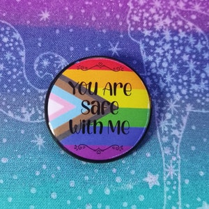 May include: A circular badge with a black edge displays the words "You Are Safe With Me" in black lettering. The design incorporates the colours of the Pride flag and the Progress Pride flag, set against a rainbow background.