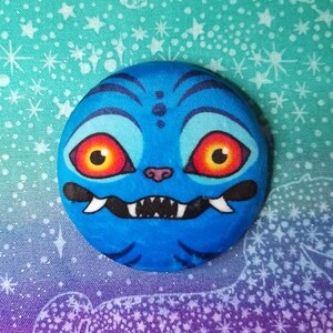 Peut inclure: Une pierre peinte bleue ronde représentant un visage de monstre de dessin animé. Le monstre a de grands yeux rouges et jaunes, un petit nez rose et une bouche noire avec des dents pointues. Le fond est un tissu turquoise avec des motifs d'étoiles blanches.