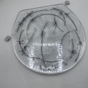 Op de afbeelding: Een heldere, ovale plaat met verschillende strengen zilverkleurig prikkeldraad over het oppervlak. Twee kleine, witte elektrische connectoren zijn zichtbaar aan de rand van de plaat. De achtergrond is wit.