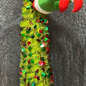 Peut inclure: Une décoration de Noël fantaisiste avec un arbre en tinsel vert citron orné de paillettes rouges et vertes. L'arbre a une paire de pieds en peluche verte avec des semelles rouges et une bordure blanche. Une boule rouge est suspendue en haut.