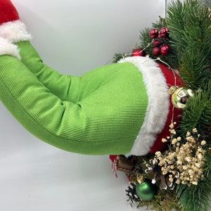 May include: A green and white Christmas wreath with a plush green elf leg sticking out of the top. The wreath is decorated with red berries, pine branches, and white flowers.