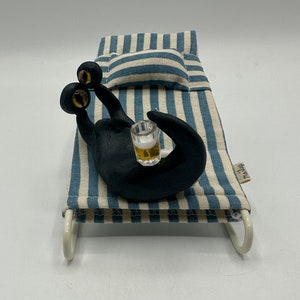 Può includere: Un divano letto in tessuto a righe bianche e nere con una creatura nera a tre occhi seduta su di esso. La creatura tiene un piccolo bicchiere di liquido. Il divano letto ha un cuscino bianco e una cornice bianca.