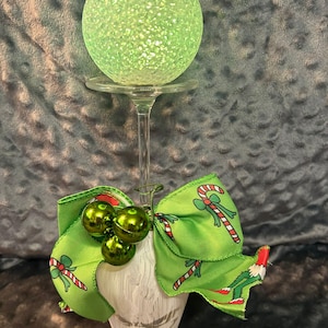 Op de afbeelding: Een decoratieve kerstversiering met een groene, gloeiende bol bovenop een heldere glazen steel. Een groene strik met zuurstok- en elf-ontwerpen siert de basis, samen met drie kleine groene balornamenten. De basis is een witte, getextureerde vaas.