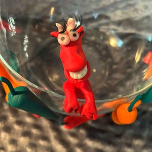 Può includere: Una stravagante statuetta di argilla rossa con grandi occhi sporgenti e lunghe ciglia, posta all'interno di una ciotola di vetro trasparente. La statuetta ha un ampio sorriso. Peperoni di argilla verde e arancione decorano il bordo della ciotola.