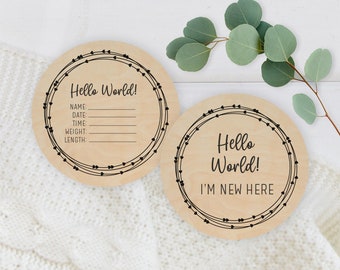 May include: Two round wooden signs with black text and a heart design. The first sign says "Hello World!" and has spaces to fill in the baby's name, date, time, weight, and length. The second sign says "Hello World! I'm New Here."