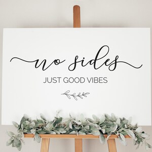 以下が含まれることがあります： 黒い書体で「no sides just good vibes」と書かれた白い看板。テキストの下に小さな黒い小枝のデザインがあります。看板はイーゼルに置かれています。