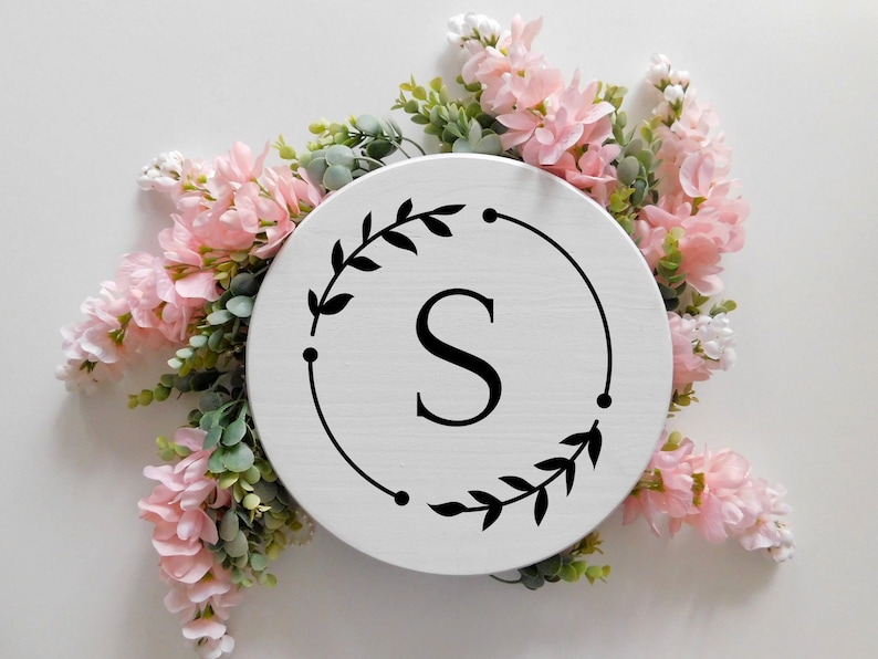 May include: White round wooden sign with a black wreath and the letter "S" in the center. The sign is surrounded by pink flowers.