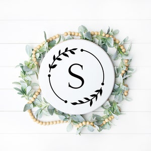 May include: White round wooden sign with a black wreath and the letter "S" in the center. The sign is surrounded by a garland of light green leaves and beige beads.