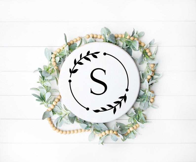 May include: White round wooden sign with a black wreath and the letter "S" in the center. The sign is surrounded by a garland of light green leaves and beige beads.