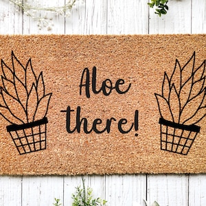 Puede incluir: Una alfombra de coco marrón con el texto "Aloe there!" en fuente cursiva negra. Dos dibujos de línea negra de plantas de aloe en maceta flanquean el texto.