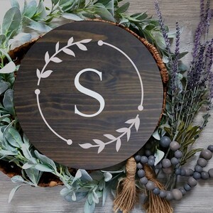 May include: A dark brown wooden round sign with a white wreath and the letter "S" in the center. The sign is surrounded by greenery and beads.