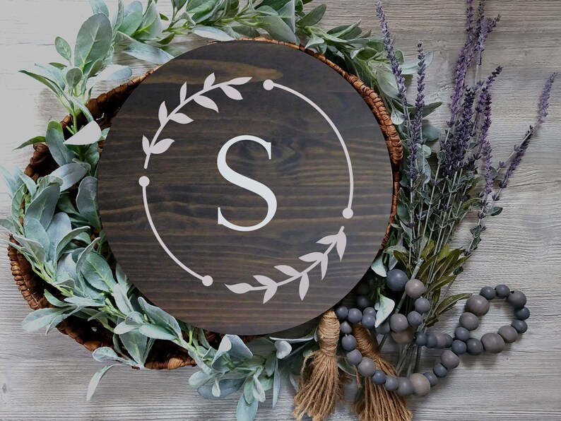 May include: A dark brown wooden round sign with a white wreath and the letter "S" in the center. The sign is surrounded by greenery and beads.