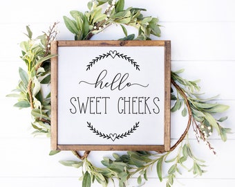 May include: A square wooden sign with the words "hello SWEET CHEEKS" in black script. The sign is surrounded by a green and white leafy garland. The sign has a brown wooden frame.