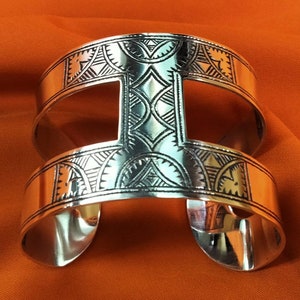 May include: Silver cuff bracelet with intricate geometric patterns. The open design features a central rectangular cutout and detailed etched lines. The bracelet is displayed on an orange background, highlighting its polished finish.