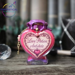May include: A heart-shaped glass bottle with a pink liquid and a purple, glittery top. The label reads "Love Potion Antidote" in a decorative font. A gold chain with beads and a key charm hangs from the bottle.