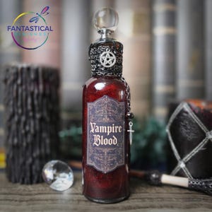 May include: A decorative glass bottle filled with red glitter and liquid, labeled "Vampire Blood." The bottle has a silver-colored chain and pentagram charm, with a clear stopper. The label is dark with ornate gold lettering.