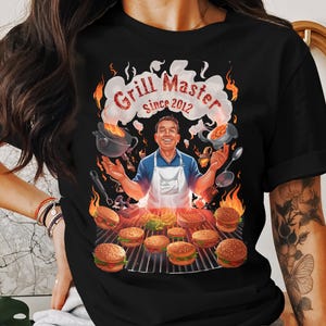 Op de afbeelding: Zwart T-shirt met een grill-illustratie. Een man met een schort is omringd door hamburgers, vlammen en kookgerei. De tekst "Grill Master since 2012" staat erop. Het ontwerp is kleurrijk en gedetailleerd.