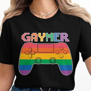 Puede incluir: Camiseta negra con la palabra "GAYMER" en letras de arcoíris pixeladas sobre un mando de videojuegos con los colores del arcoíris. El mando tiene una cruceta, botones y palancas analógicas.